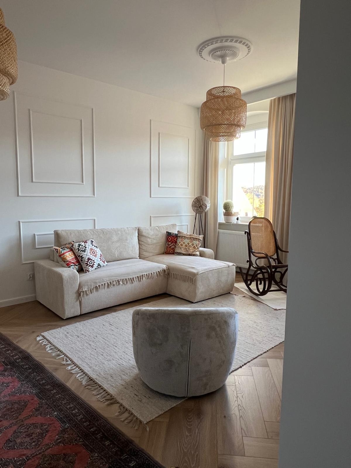 Jasne wnętrze salonu z beżową sofą narożną, dekoracyjnymi listwami na ścianach, dywanem i wiklinową lampą. Widoczne elementy wykończenia wnętrz.