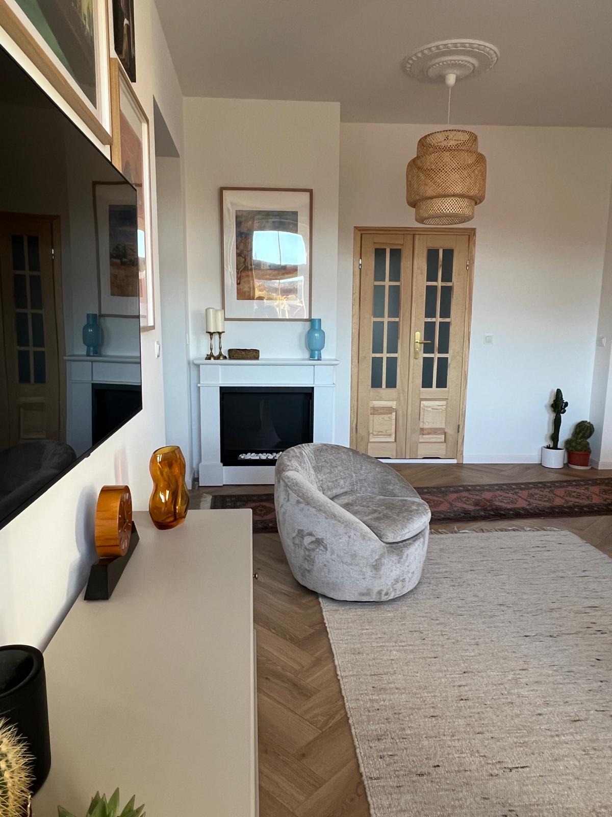 Jasne wnętrze salonu z parkietem w jodełkę, kremowym dywanem i fotelem typu muszelka. Kominek z dekoracjami i drewniane drzwi dwuskrzydłowe.