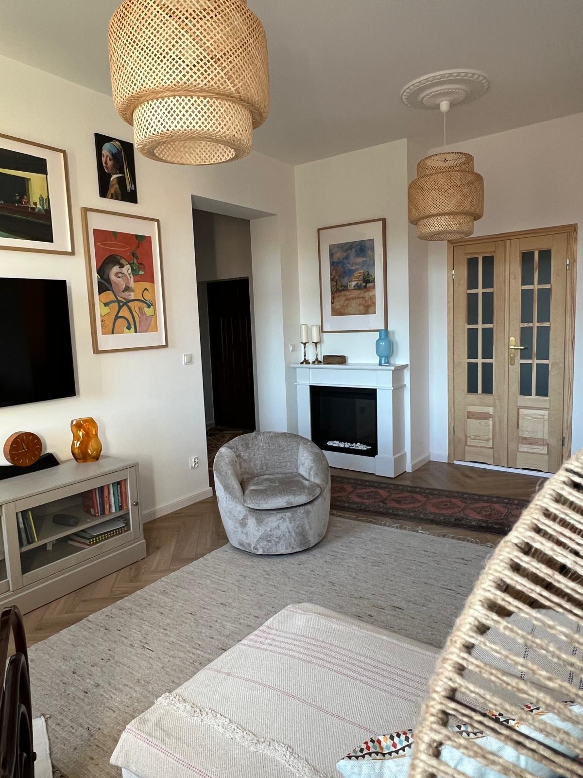 Jasne wnętrze salonu z drewnianą podłogą, kominkiem i rattanowymi lampami. Na ścianie wiszą obrazy. Widoczne drzwi z jasnego drewna.