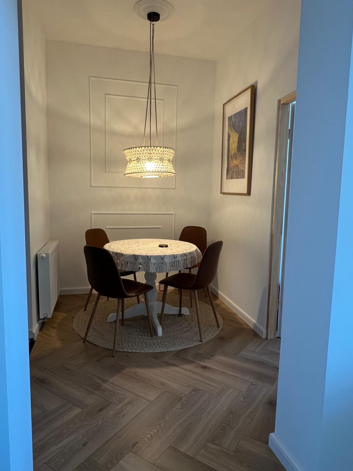 Jasne wnętrze jadalni z okrągłym stołem, czterema krzesłami i ozdobną lampą. Na ścianie dekoracyjny panel i obraz. Parkiet ułożony w jodełkę. Całość w neutralnej tonacji.