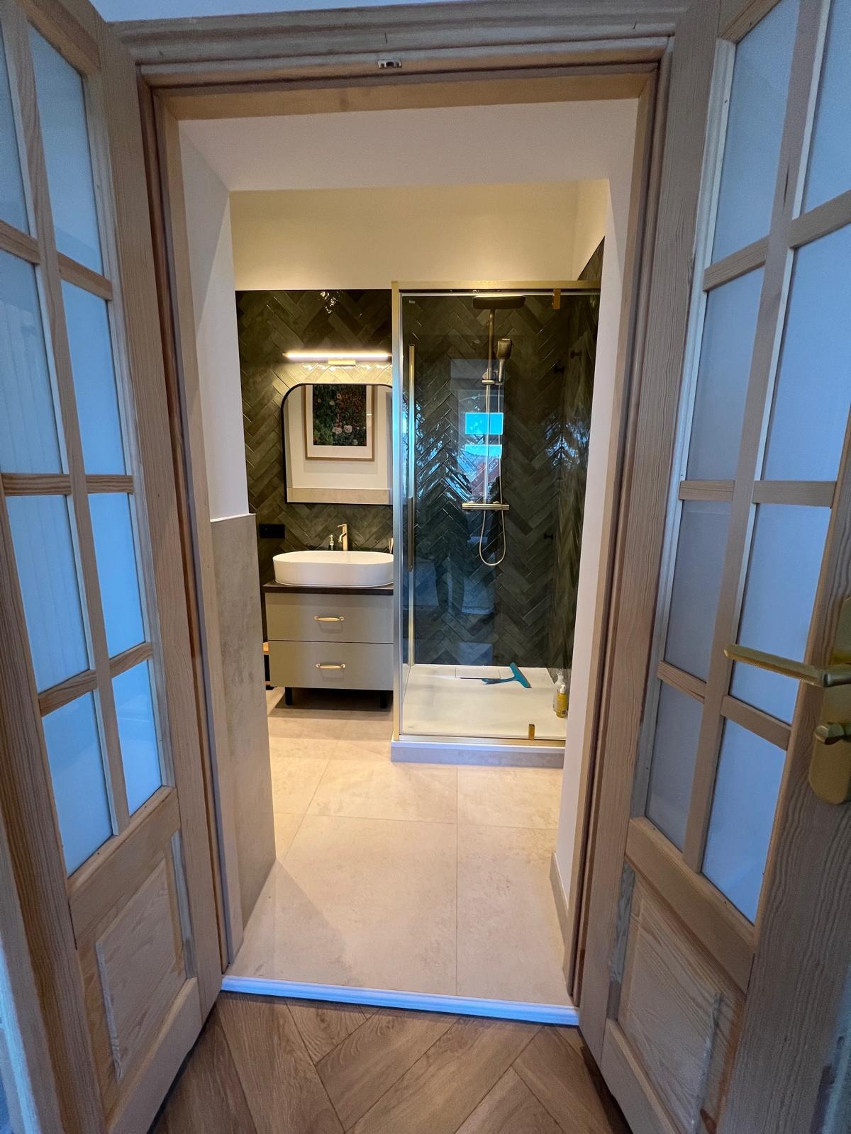 Nowoczesna łazienka z kabiną prysznicową w złotej ramie, umywalką na szafce i lustrem, widziana przez otwarte, drewniane drzwi.