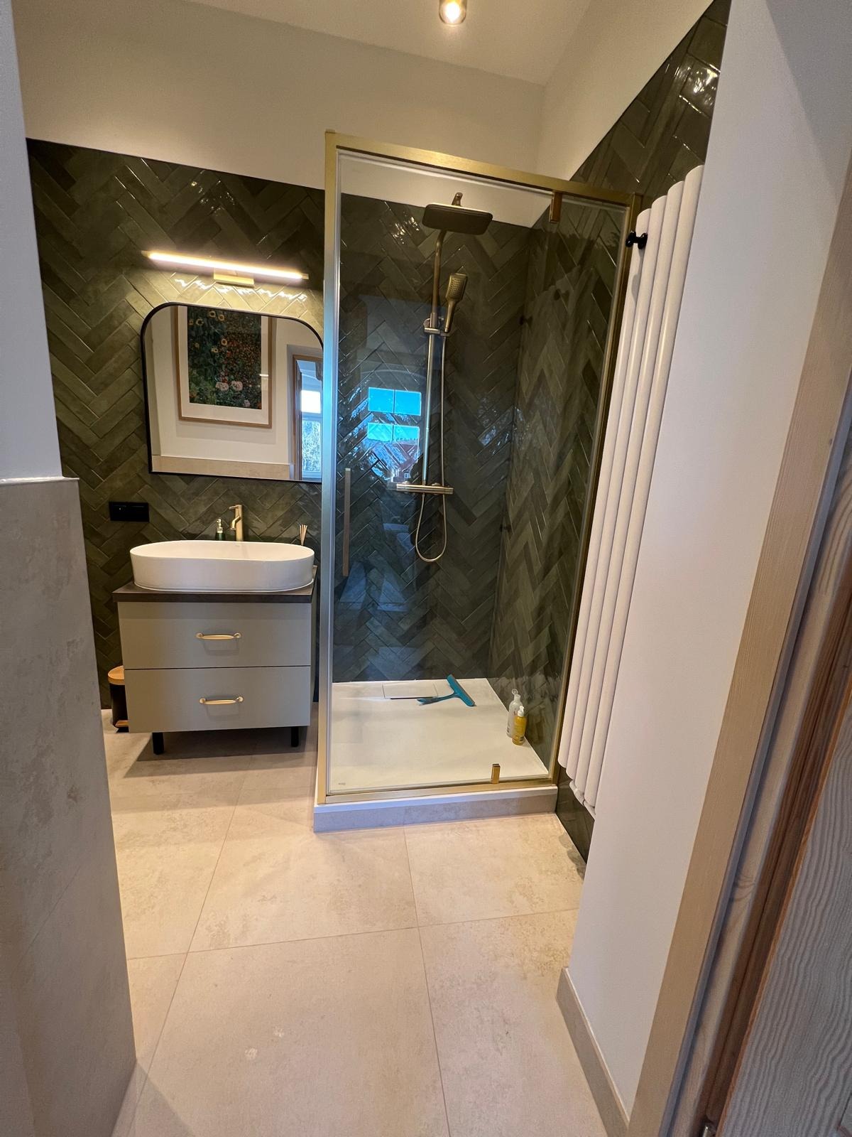 Stylowa łazienka z kabiną prysznicową w złotej ramie i umywalką na szafce. Ściany wyłożone zieloną, wzorzystą glazurą w jodełkę. Uwagę przykuwa lustro z oświetleniem.