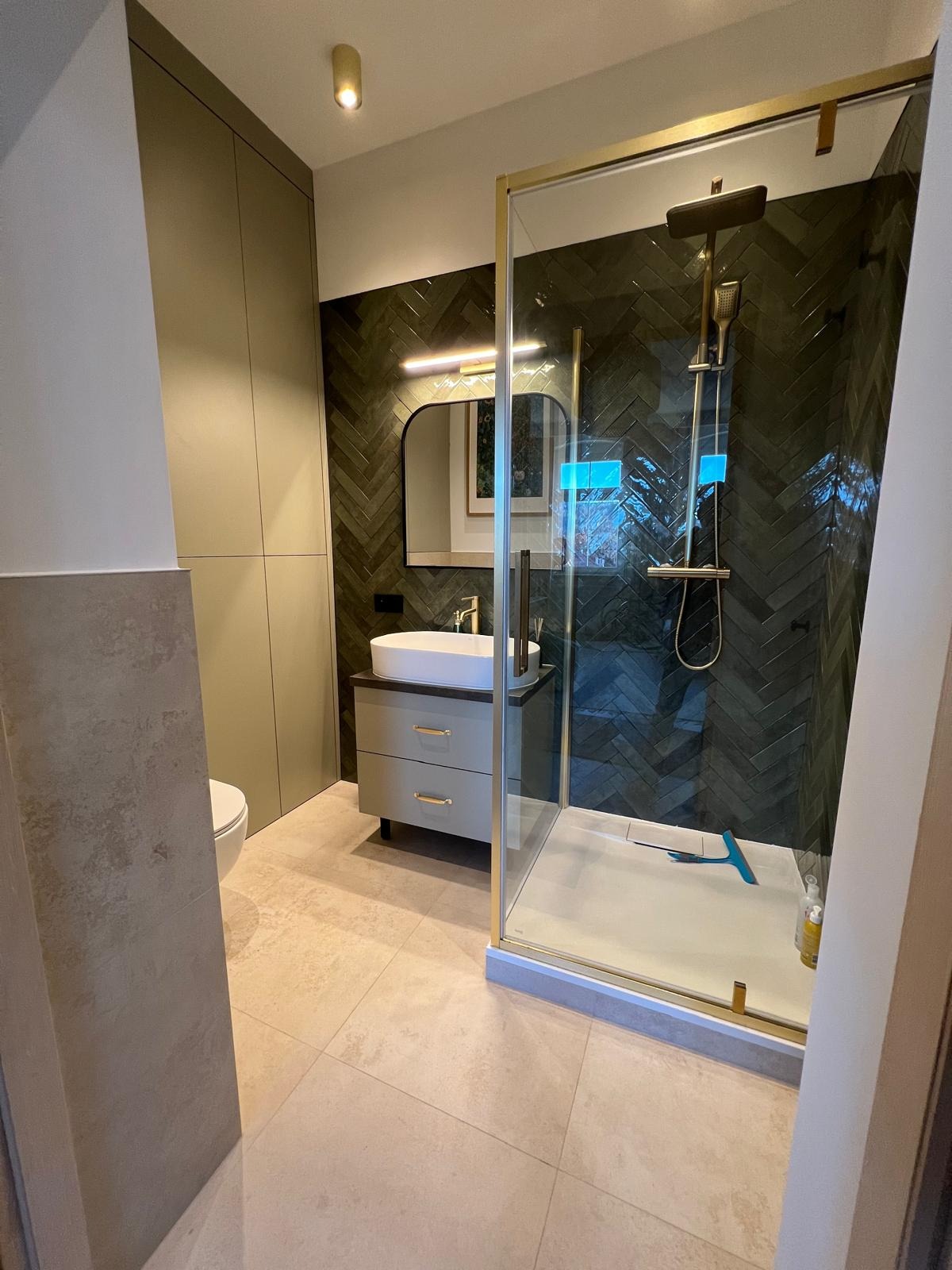 Nowoczesna łazienka z kabiną prysznicową w złotej ramie, umywalką na szafce i ścianą z ciemnej, ułożonej w jodełkę glazury. Minimalistyczne oświetlenie.