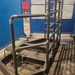 Konstrukcja metalowa ze schodami i barierkami, wykonana z rur i profili, spawana, widoczne spawy. W tle niebieskie ekrany i rysunki techniczne.