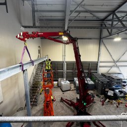 S&M Stanisław Maślanka - Spawacz w kasku, na podnośniku Hoeflon, montuje stalową belkę w hali przemysłowej. Widok z góry, dźwig, kontenery, schody.