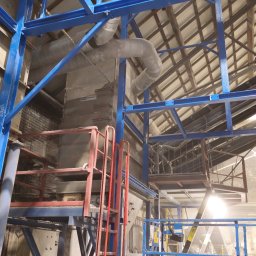 S&M Stanisław Maślanka - Konstrukcja stalowa z rurami i podnośnikiem w hali produkcyjnej w Markuszowej. Niebieska stalowa rama i czerwona drabina. Widok na sufit hali.