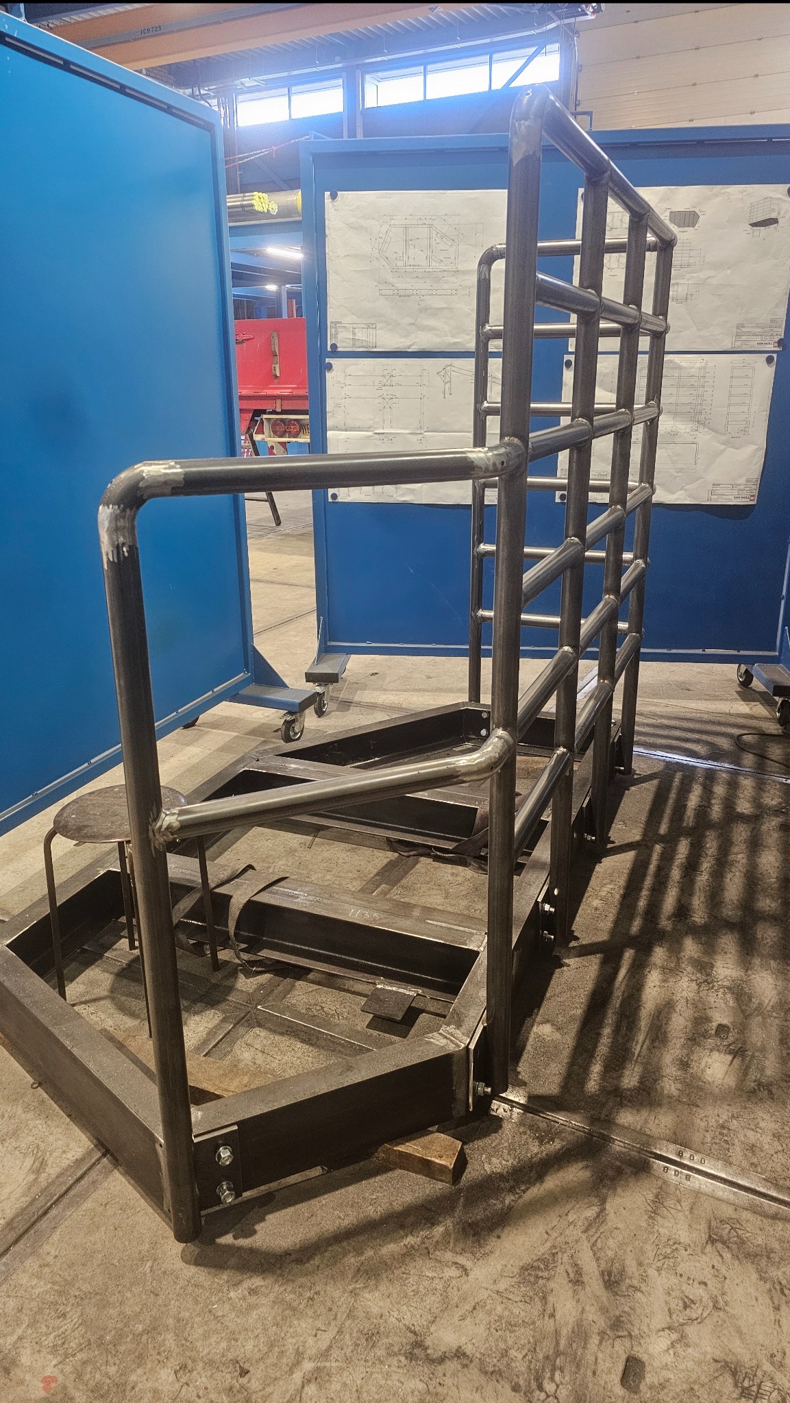 Konstrukcja metalowa ze schodami i barierkami, wykonana z rur i profili, spawana, widoczne spawy. W tle niebieskie ekrany i rysunki techniczne.