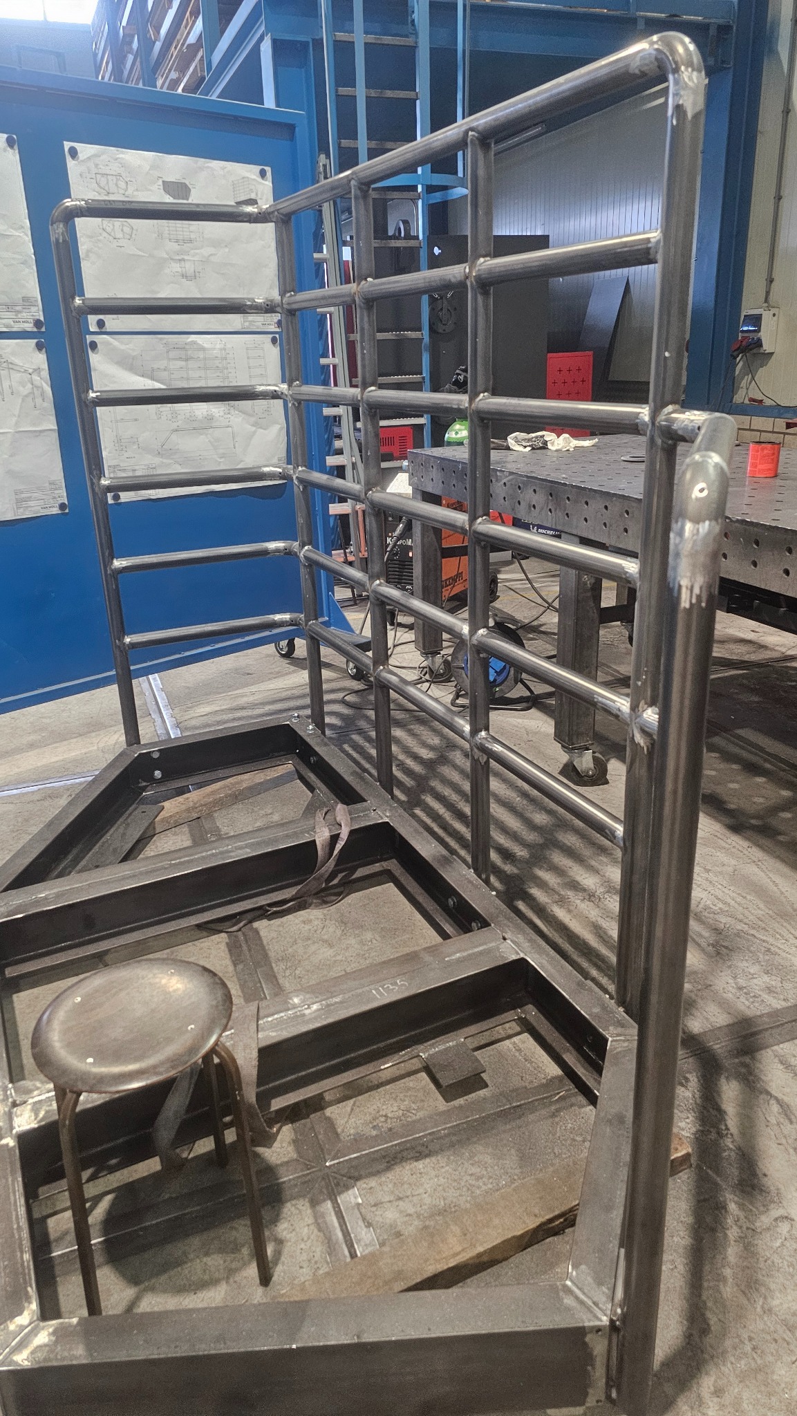 Stalowa konstrukcja z rur i profili, spawana na warsztacie. Widoczny stół spawalniczy w tle i krzesło obok konstrukcji. Elementy konstrukcyjne z widocznymi spawami.