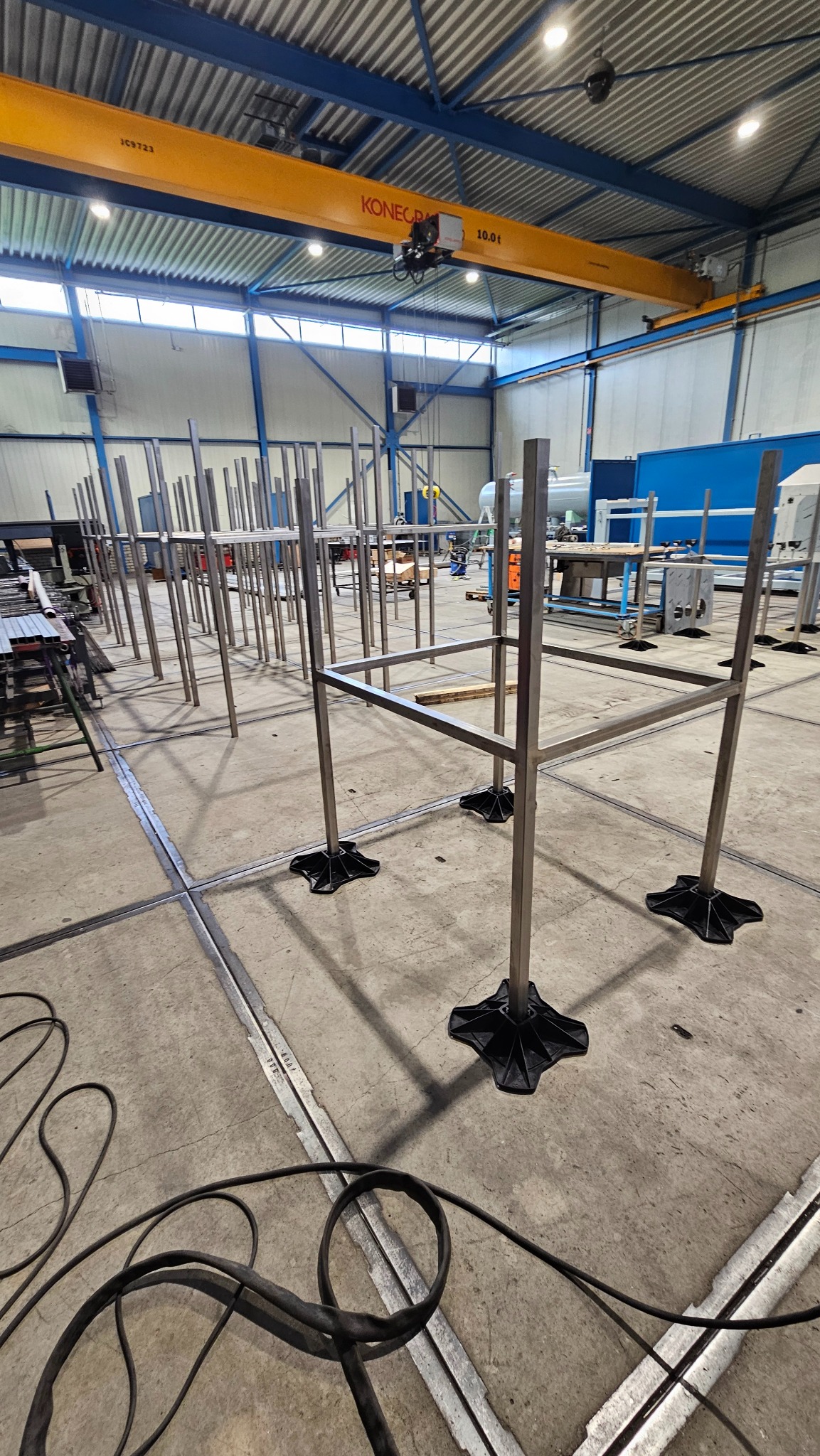 Konstrukcja metalowa z profili na czarnych podstawach w hali produkcyjnej z suwnicą. Widoczne kable na podłodze i inne elementy wyposażenia hali.