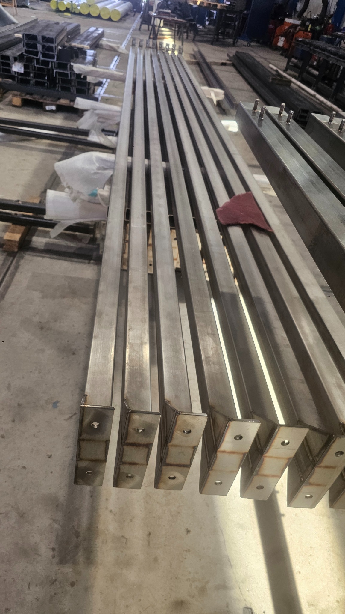 Rzędy metalowych profili z przyspawanymi elementami mocującymi, ułożone równolegle w warsztacie. Widoczne ślady po obróbce i spawaniu.