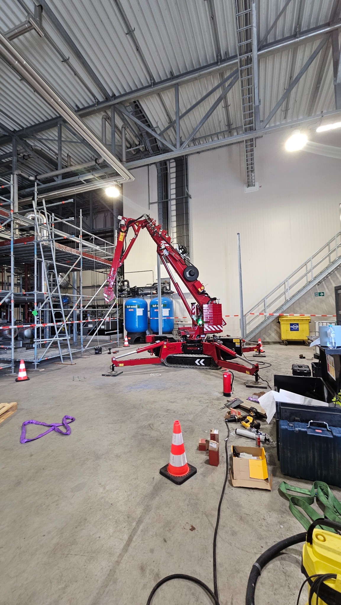 Wnętrze hali produkcyjnej z suwnicą gąsienicową do prac montażowych. Widoczne elementy instalacji, rusztowania, narzędzia i zabezpieczenia budowlane.