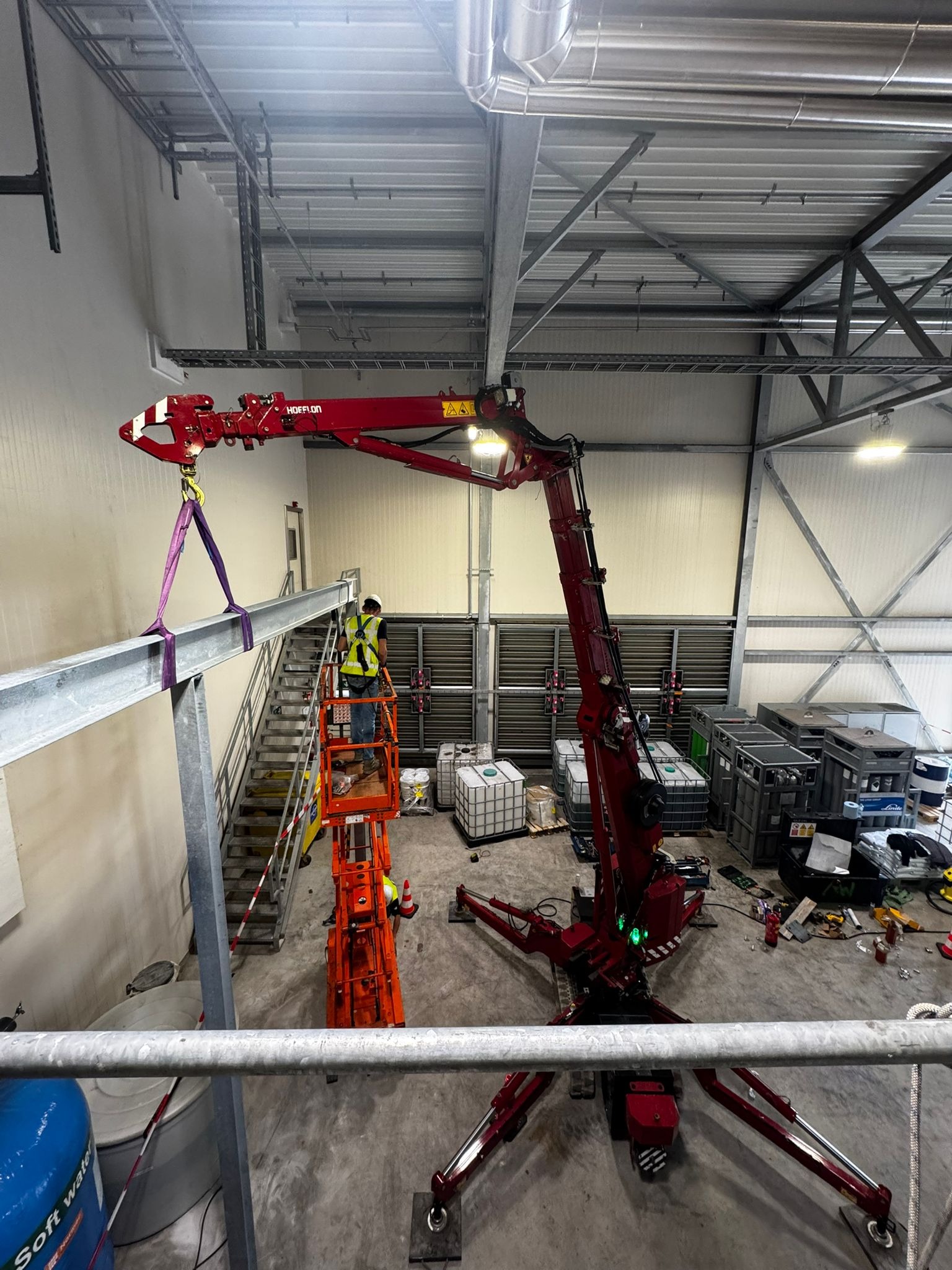 Spawacz w kasku, na podnośniku Hoeflon, montuje stalową belkę w hali przemysłowej. Widok z góry, dźwig, kontenery, schody.