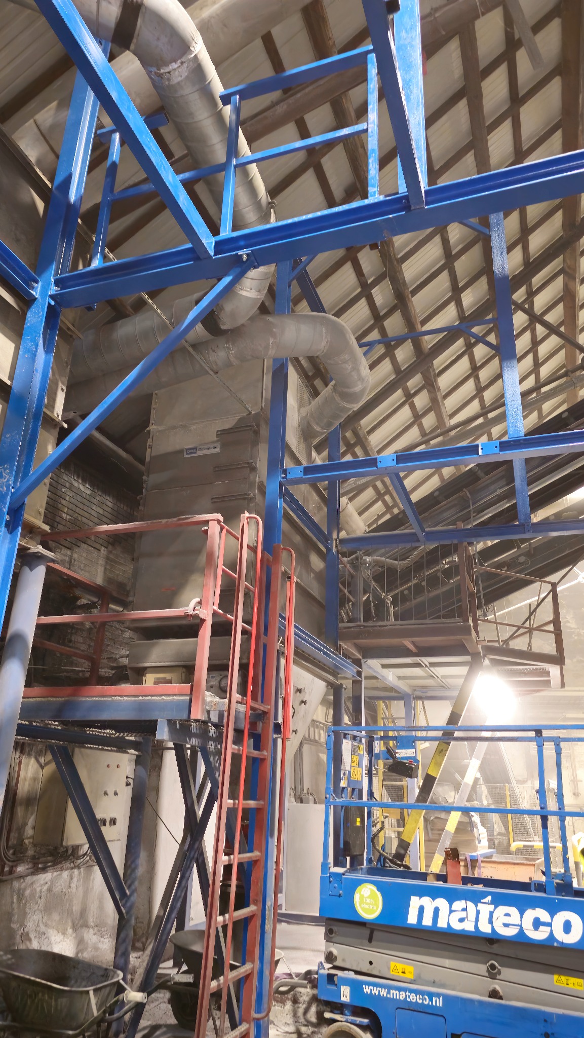 Konstrukcja stalowa z rurami i podnośnikiem w hali produkcyjnej w Markuszowej. Niebieska stalowa rama i czerwona drabina. Widok na sufit hali.