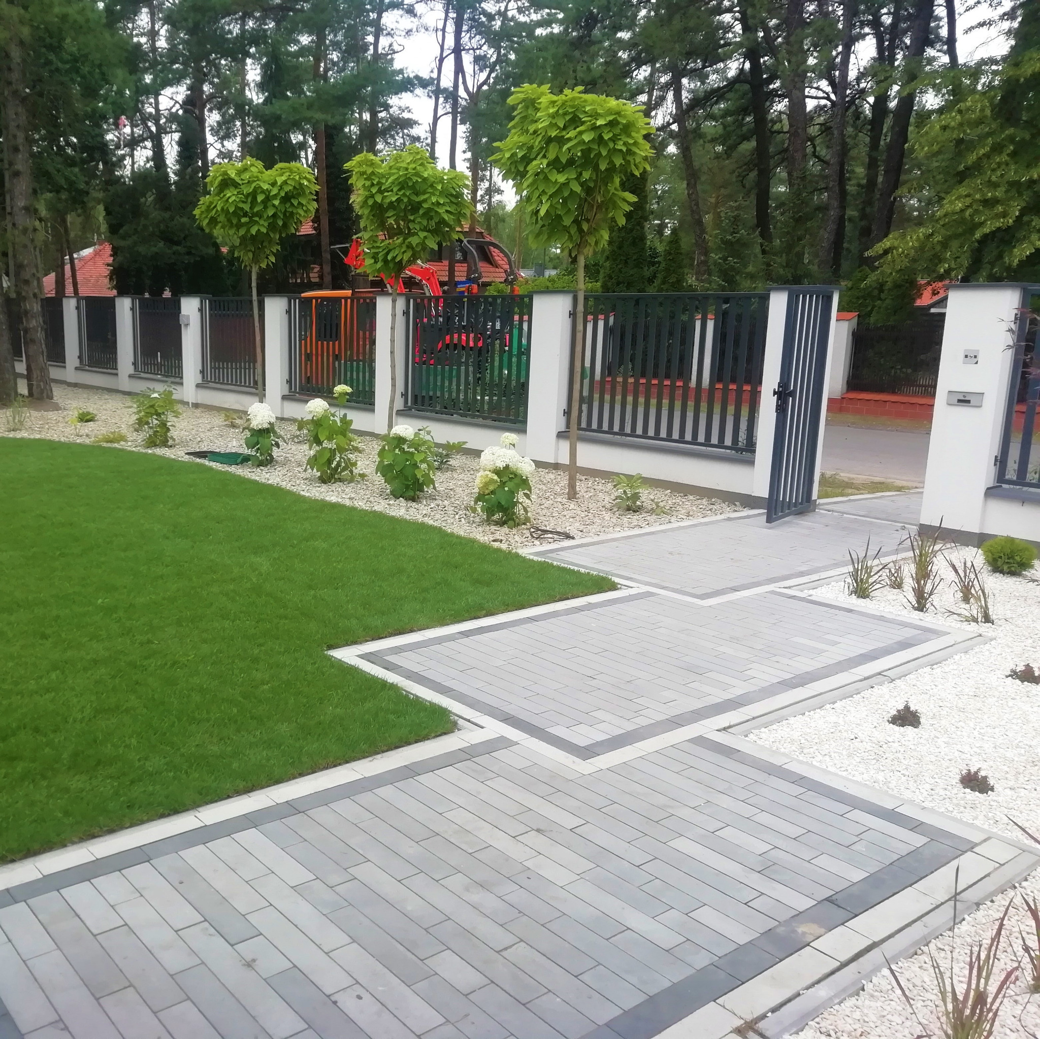 Nowoczesny ogród z równo przystrzyżonym trawnikiem, ścieżką z kostki brukowej i ozdobnymi krzewami na tle ogrodzenia i drzew. Widoczne elementy placu zabaw.