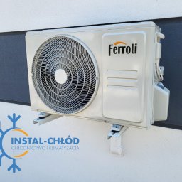 INSTAL-CHŁÓD CHŁODNICTWO-KLIMATYZACJA DAWID JEWASIŃSKI - Klimatyzator Ferroli zamontowany na białej elewacji budynku w Łabiszynie. Widoczne mocowania i fragment logo firmy Instal-Chłód na dole zdjęcia.