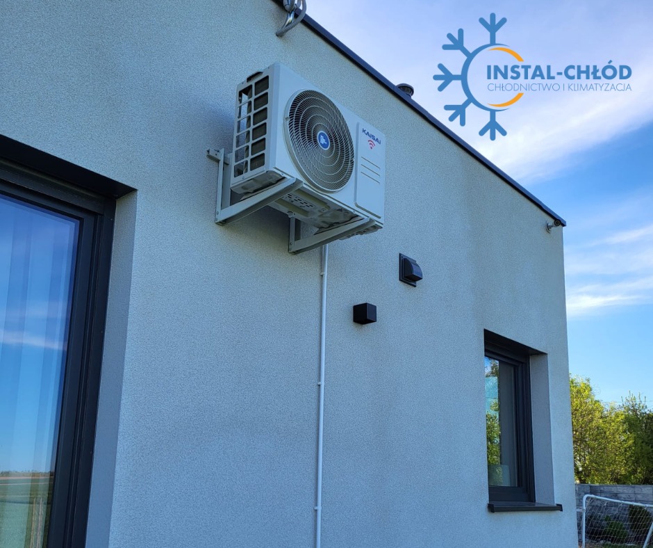 Biała klimatyzacja KANSAI zamontowana na elewacji domu z widocznymi oknami i błękitnym niebem w tle. Instalacja wykonana przez INSTAL-CHŁÓD.
