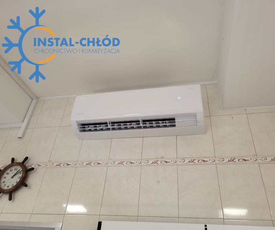 Biały klimatyzator ścienny z wyświetlaczem temperatury, zamontowany na kafelkowanej ścianie. Widoczny zegar w kształcie steru i logo firmy w rogu.