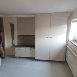 Meble & Pasją - Jasna zabudowa przedpokoju z szafą, siedziskiem i wieszakami. Uzupełnieniem jest grzejnik pod oknem i płytki podłogowe w szarym odcieniu.