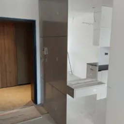 Montaż szafy z lustrzanymi frontami w przedpokoju; widoczne zabezpieczenia folią. W tle fragment kuchni i drzwi do pokoju. Nowoczesne wykończenie wnętrza.