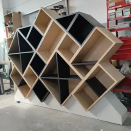 Nietypowa, geometryczna szafka z drewnianymi i czarnymi elementami, ustawiona w warsztacie. Widoczne narzędzia i regał z akcesoriami w tle.