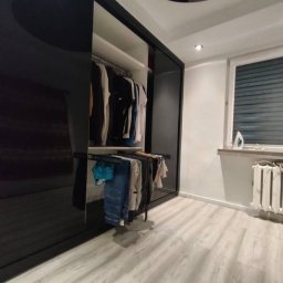 Meble & Pasją - Nowoczesna szafa wnękowa z przesuwnymi, czarnymi drzwiami na wysoki połysk. Wnętrze szafy z ubraniami na wieszakach i wysuwanym drążku.