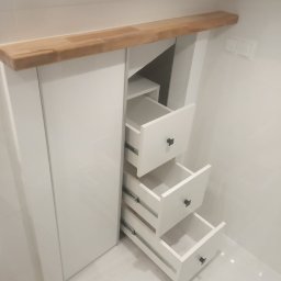 Meble & Pasją - Biała szafa wnękowa z trzema wysuniętymi szufladami i drewnianym blatem, wkomponowana w białe, kafelkowane wnętrze. Widoczne metalowe prowadnice szuflad.