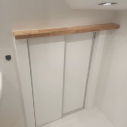 Meble & Pasją - Biała szafa wnękowa z przesuwnymi drzwiami i drewnianą półką nad nią, w minimalistycznej łazience. Ujęcie z dołu, widoczny fragment podłogi.