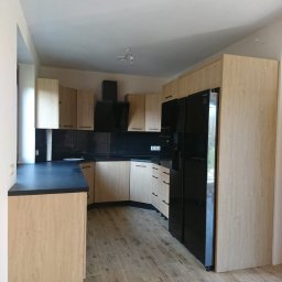 Meble & Pasją - Jasna kuchnia w kształcie litery L z czarnym blatem i sprzętem AGD. Fronty szafek z delikatnym wzorem drewna. Uwagę przykuwają minimalistyczne uchwyty.