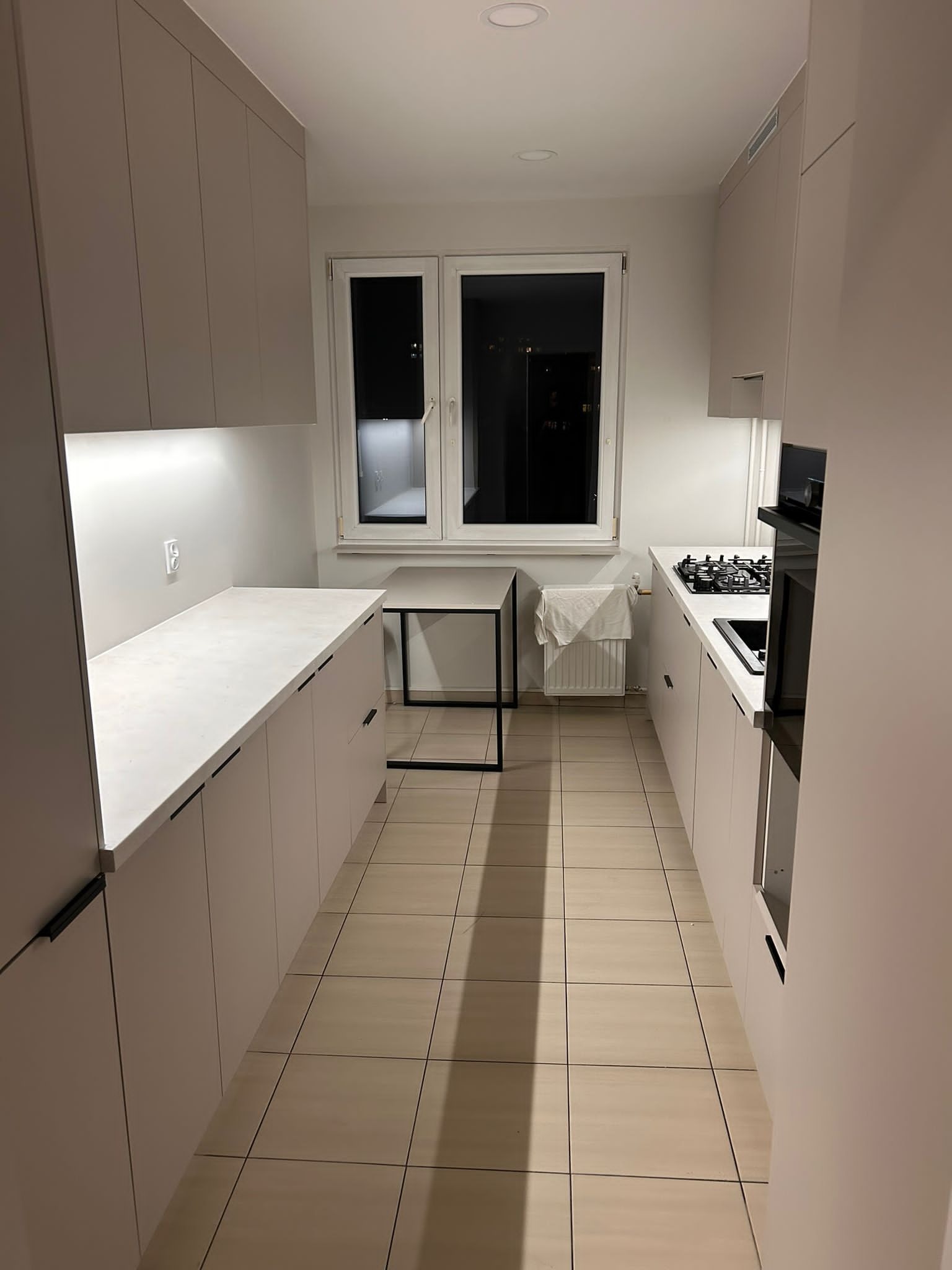 Wąska kuchnia w jasnych barwach z białym blatem i czarnymi uchwytami. Widok na okno z widokiem na nocne miasto. Minimalistyczny stół z czarnymi nogami.