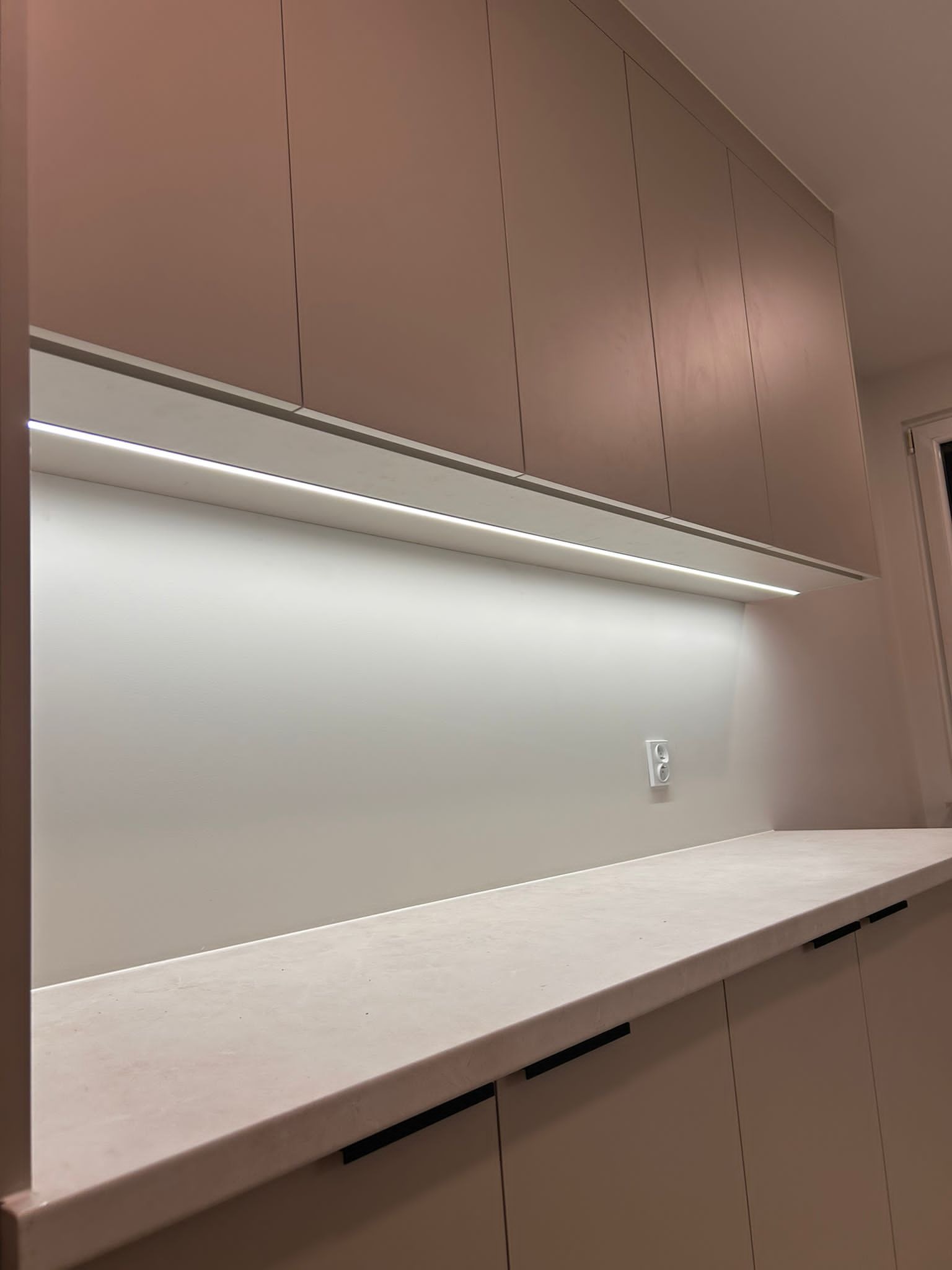 Jasna kuchnia z oświetleniem LED podszafkowym i minimalistycznymi uchwytami. Widoczny fragment blatu i gniazdko elektryczne w tle.