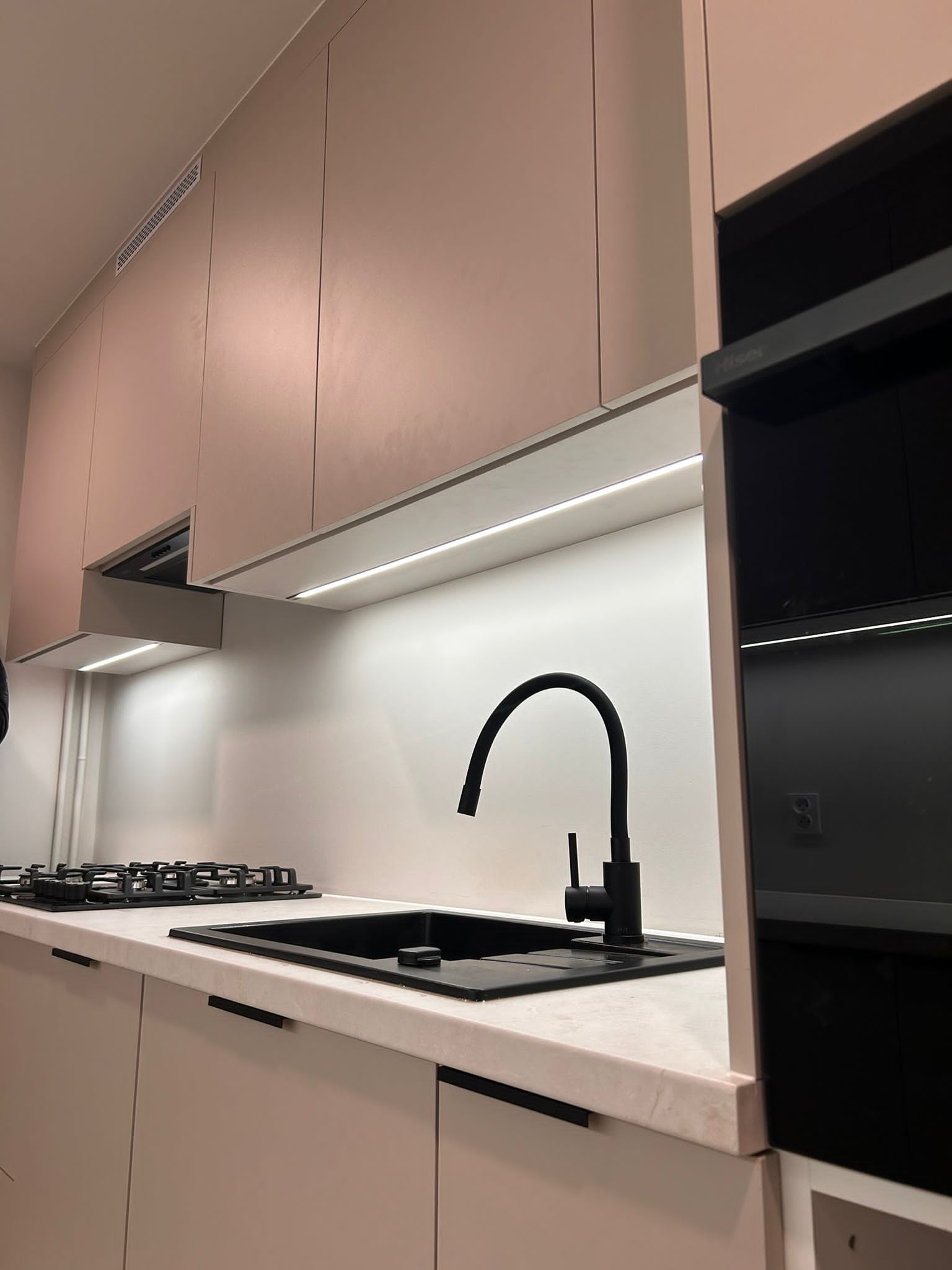 Minimalistyczna kuchnia: Jasne fronty szafek, czarny zlew i bateria, oświetlenie LED pod szafkami wiszącymi, płyta gazowa. Nowoczesny design.