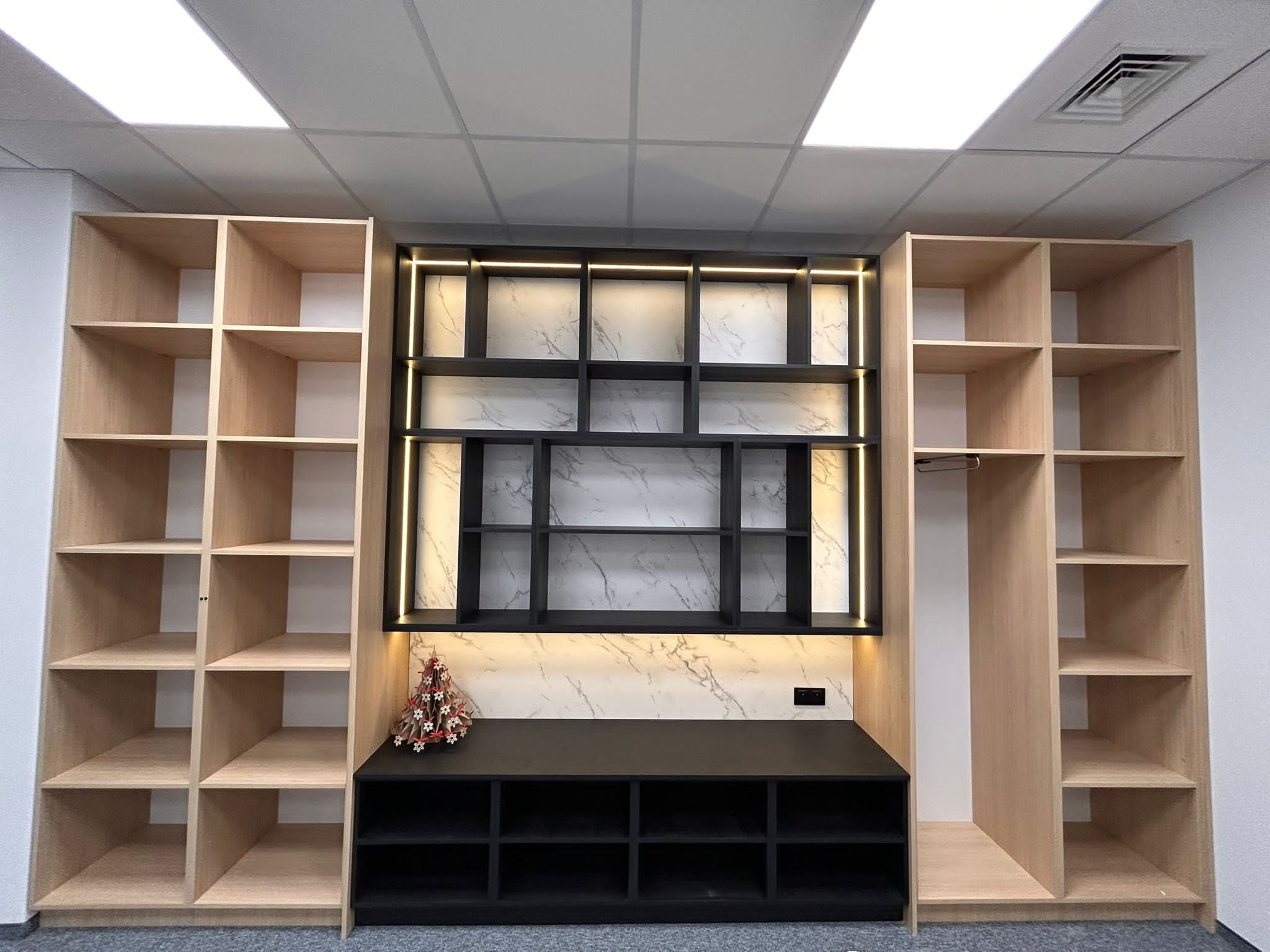 Zabudowa wnęki z jasnego drewna z otwartymi półkami, czarnym blatem i podświetlanymi elementami na marmurowym tle. Dekoracja świąteczna na blacie.