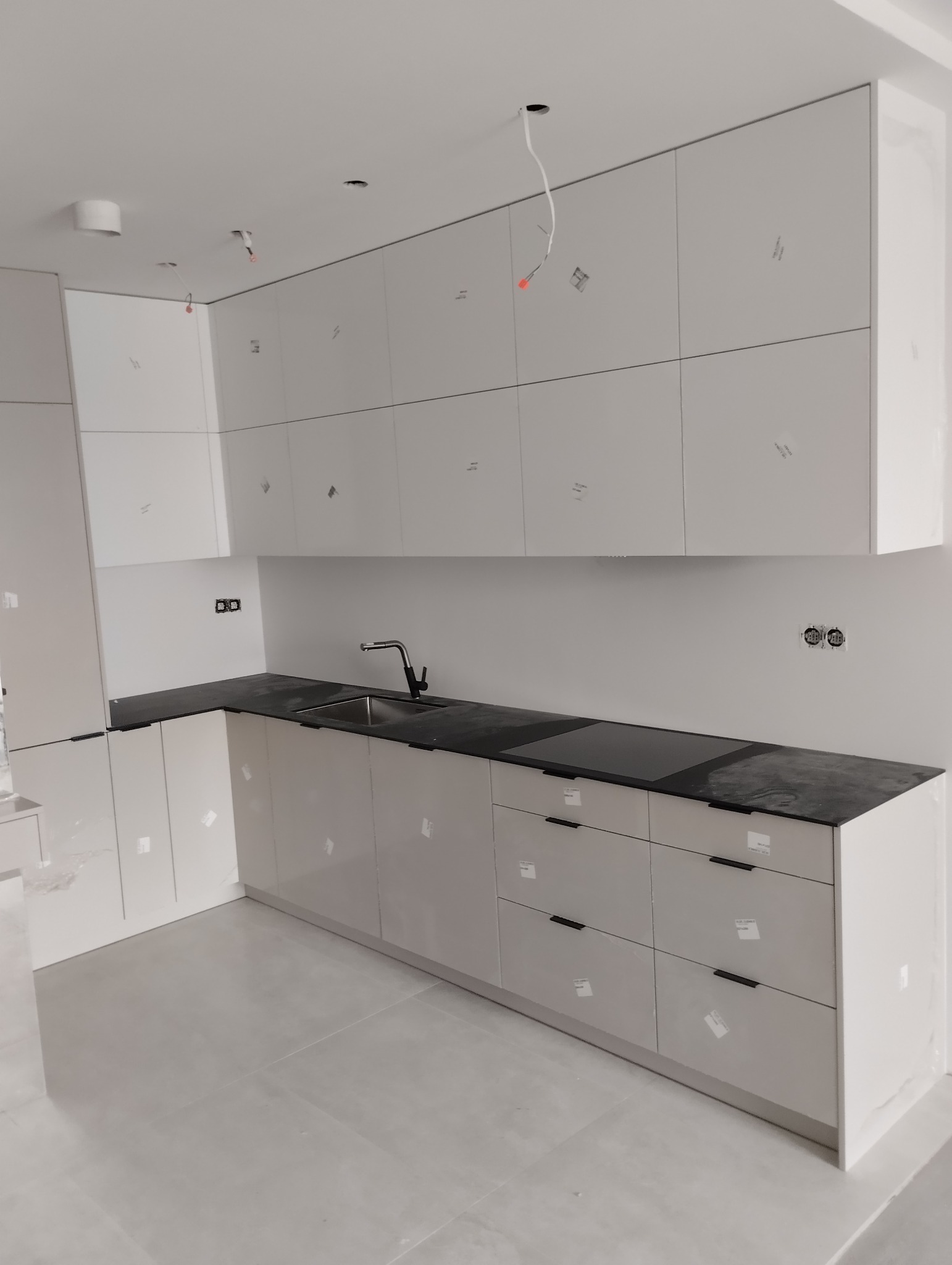 Nowoczesna kuchnia z szarymi, lakierowanymi frontami i czarnym blatem. Widoczne niedokończone prace wykończeniowe: wystające kable z sufitu.