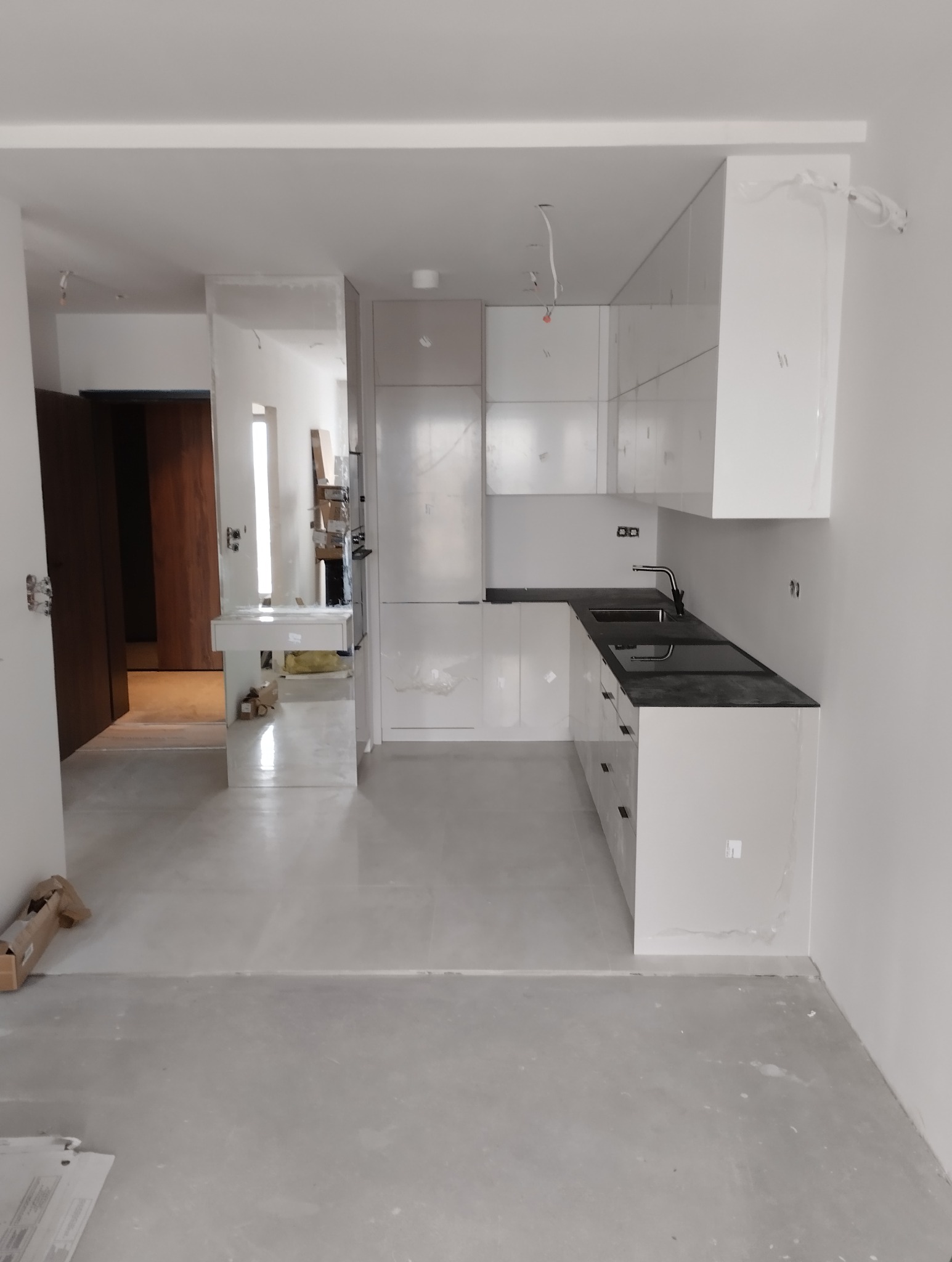 Jasna kuchnia z czarnym blatem w surowym wnętrzu. Widoczne niedociągnięcia wykończeniowe i brak listew przypodłogowych. Minimalistyczny design, otwarte przejście do dalszej części domu.