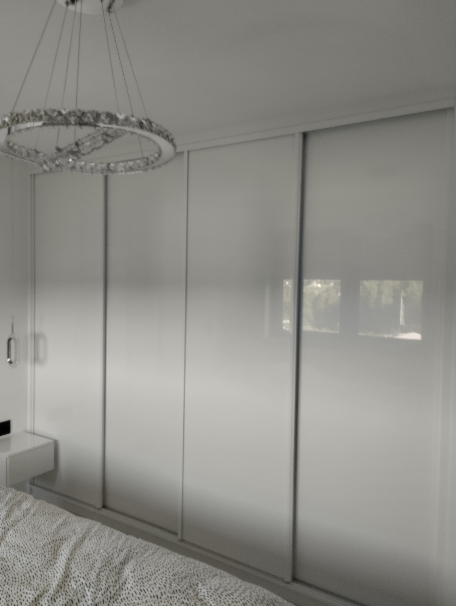 Nowoczesna szafa wnękowa z przesuwnymi, białymi drzwiami, oświetlona designerskim żyrandolem w sypialni. Minimalistyczny styl.