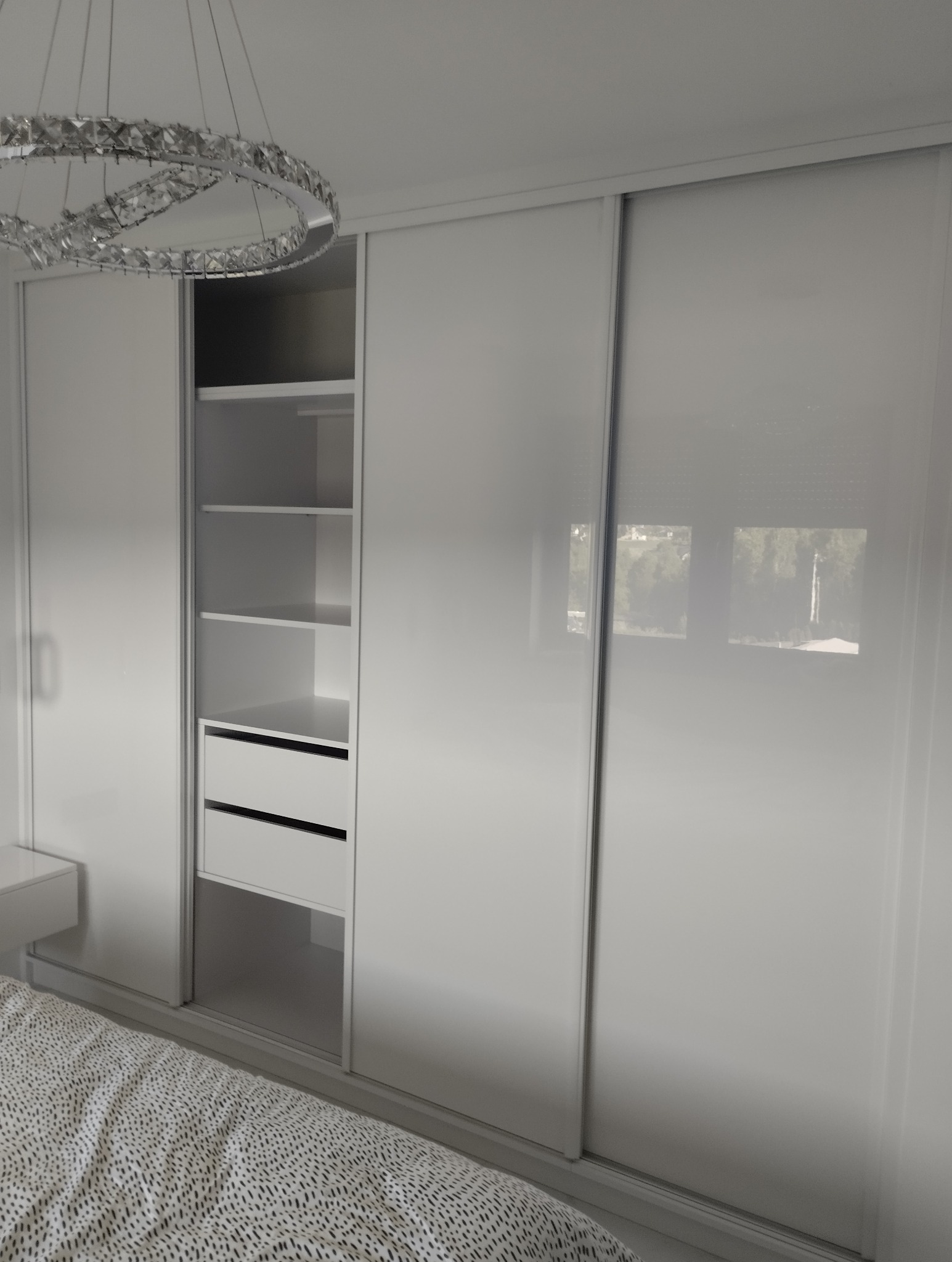 Biała szafa przesuwna z otwartą częścią z półkami i szufladami, oświetlona ozdobnym żyrandolem. Minimalistyczny design w sypialni.