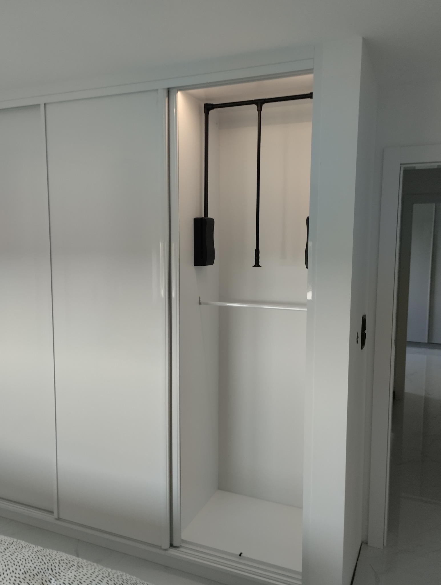 Biała szafa wnękowa z przesuwnymi drzwiami, widoczne wnętrze z drążkiem i pantografem. Minimalistyczny design, jasne oświetlenie, widok na wnętrze szafy.