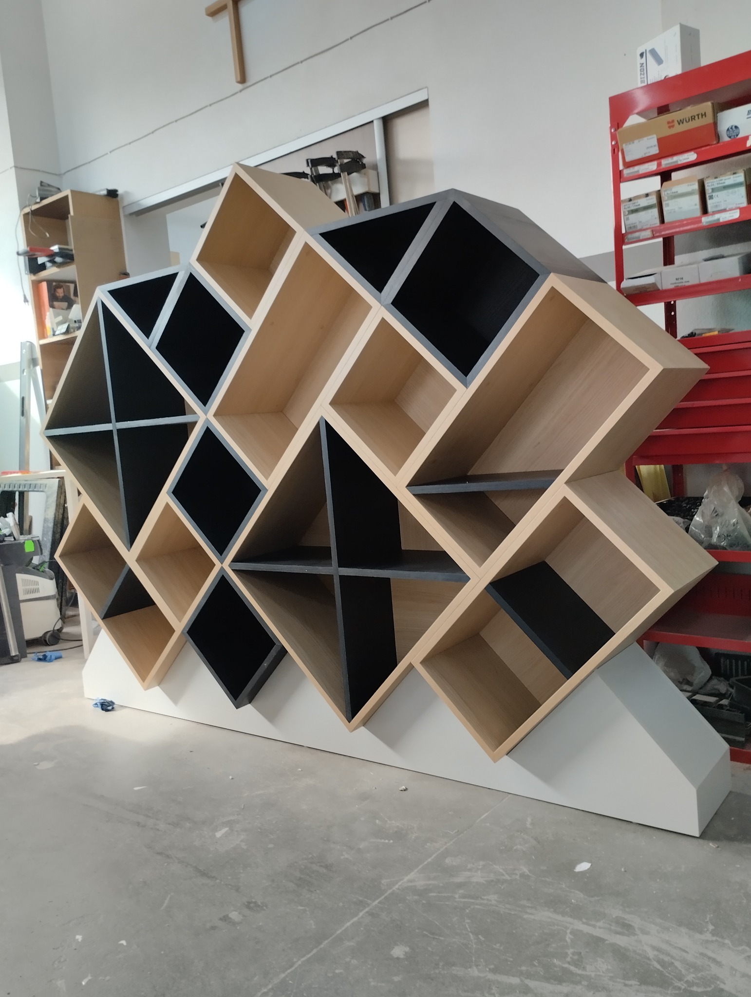 Nietypowa, geometryczna szafka z drewnianymi i czarnymi elementami, ustawiona w warsztacie. Widoczne narzędzia i regał z akcesoriami w tle.