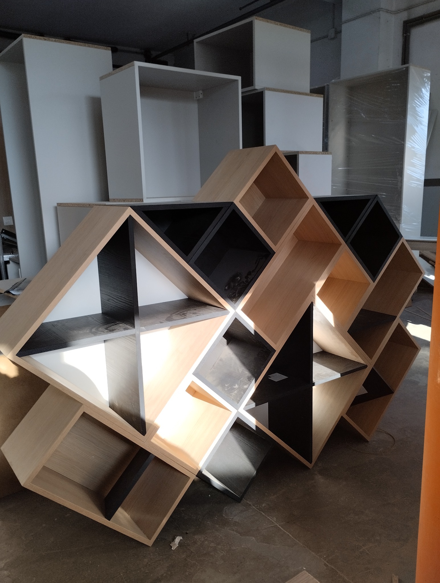Nietypowa, geometryczna konstrukcja z modułów szafek w odcieniach drewna i czerni, w tle białe korpusy szafek, surowy stan montażu.
