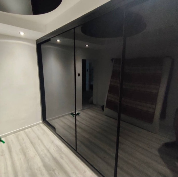 Nowoczesna szafa wnękowa z czarnymi, lustrzanymi drzwiami przesuwnymi, odbijającymi wnętrze pokoju. Widoczny fragment podłogi z paneli i oświetlenie sufitowe.