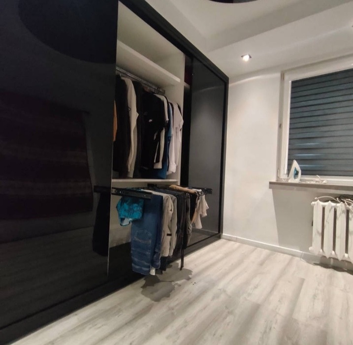 Nowoczesna szafa wnękowa z przesuwnymi, czarnymi drzwiami na wysoki połysk. Wnętrze szafy z ubraniami na wieszakach i wysuwanym drążku.