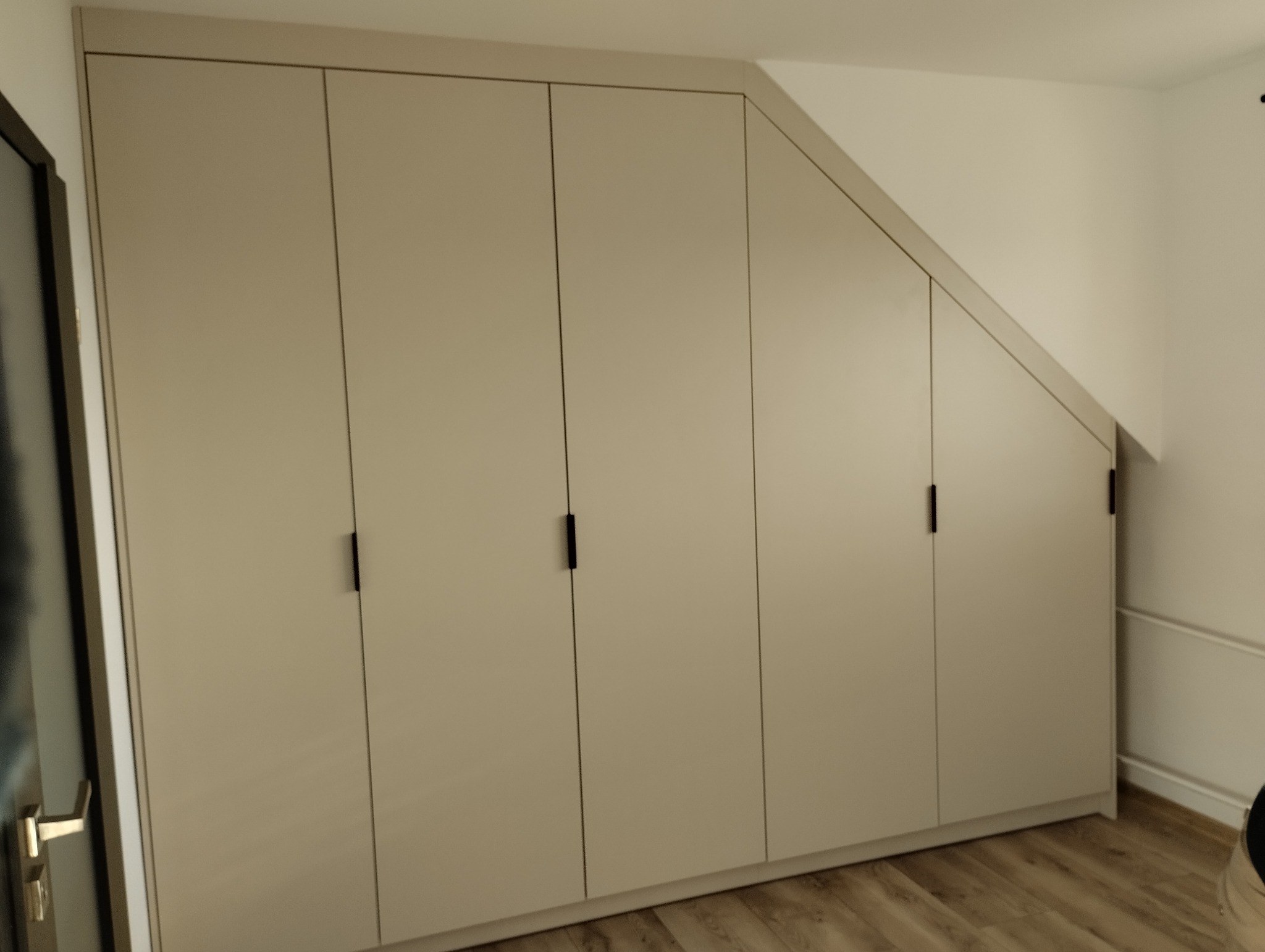Beżowa szafa wnękowa z czarnymi uchwytami, dopasowana do skosu dachu, na drewnianej podłodze. Minimalistyczny design, jasne wnętrze.