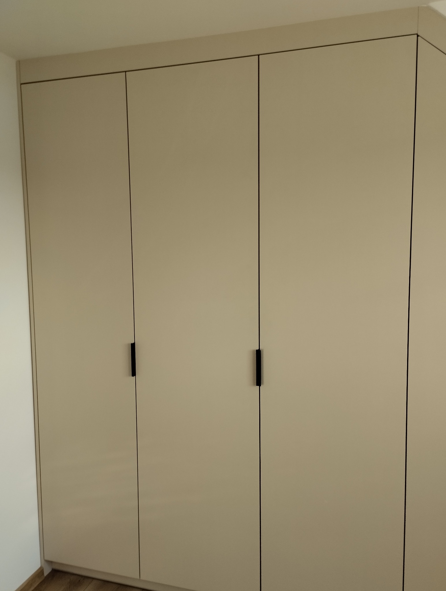 Jasna szafa trzydrzwiowa z minimalistycznymi, czarnymi uchwytami, dopasowana do wnętrza. Proste linie i neutralna kolorystyka.