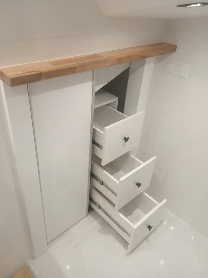 Biała szafa wnękowa z trzema wysuniętymi szufladami i drewnianym blatem, wkomponowana w białe, kafelkowane wnętrze. Widoczne metalowe prowadnice szuflad.