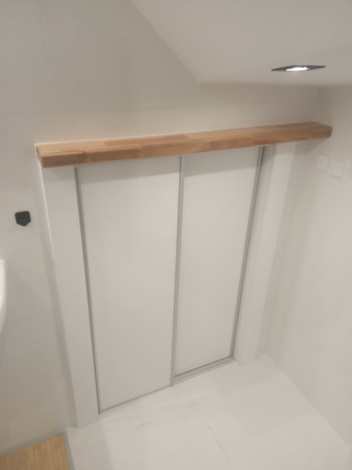 Biała szafa wnękowa z przesuwnymi drzwiami i drewnianą półką nad nią, w minimalistycznej łazience. Ujęcie z dołu, widoczny fragment podłogi.