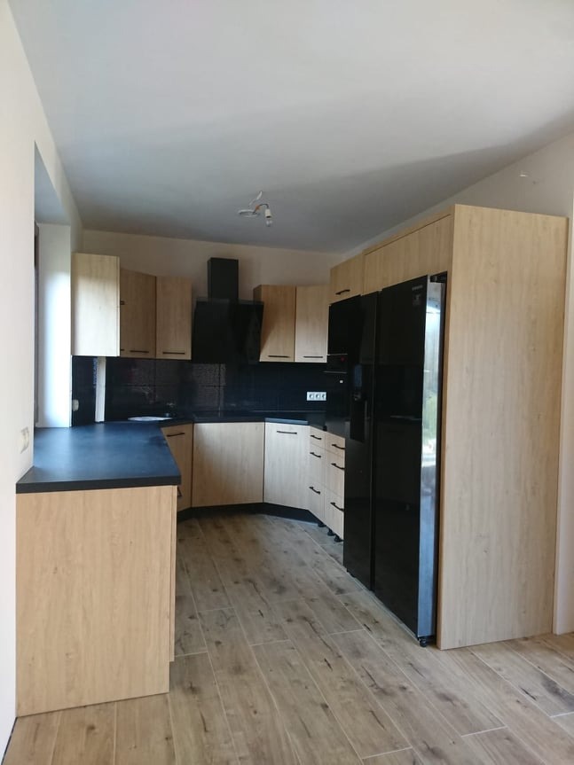 Jasna kuchnia w kształcie litery L z czarnym blatem i sprzętem AGD. Fronty szafek z delikatnym wzorem drewna. Uwagę przykuwają minimalistyczne uchwyty.