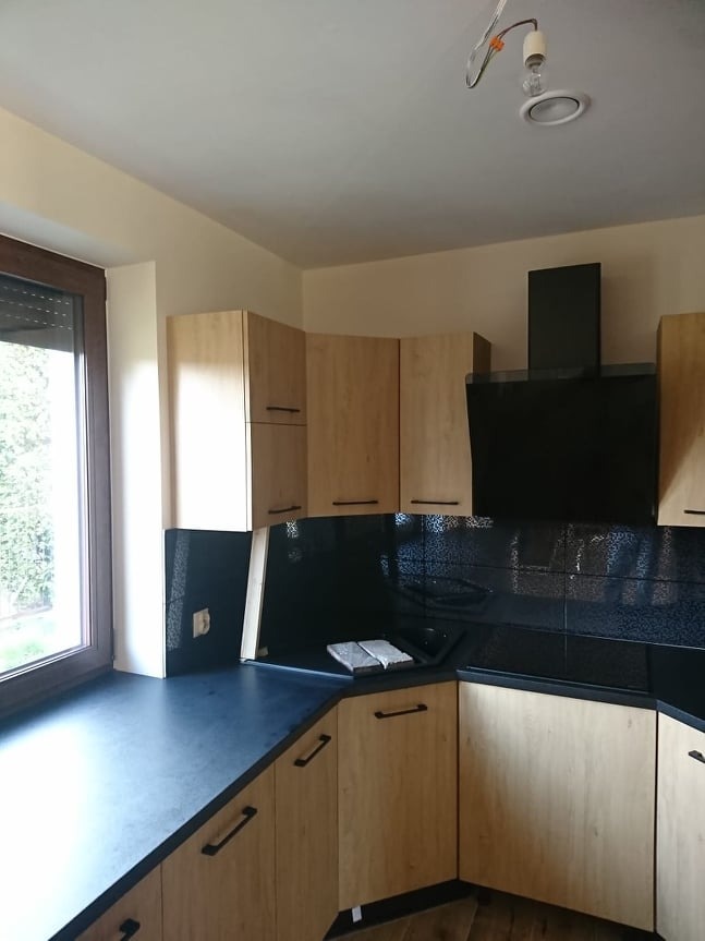 Jasna, narożna kuchnia z czarnym blatem i mozaiką nad szafkami. Widoczny okap i okno z roletami. W tle sufit z lampą bez klosza. Ujęcie z dołu.