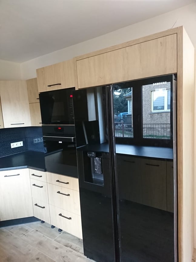 Jasna kuchnia z czarnym blatem i sprzętem AGD. Uwagę przykuwa lodówka side-by-side z dozownikiem wody i kostkarką. Minimalistyczne uchwyty szafek.