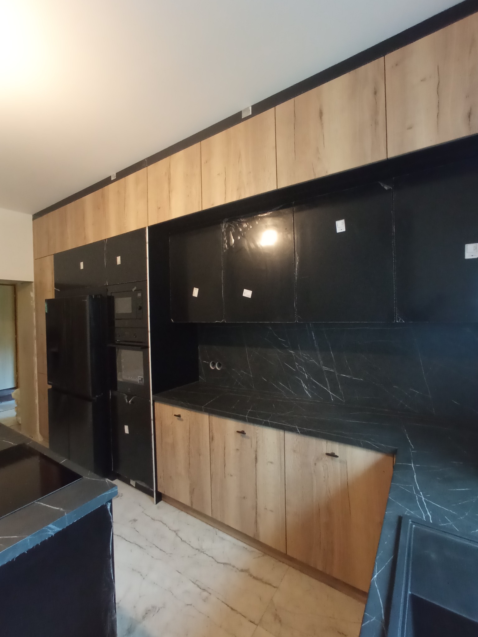 Nowoczesna kuchnia z drewnianymi szafkami i czarnym blatem w Rybniku. Widoczny sprzęt AGD w zabudowie i marmurowe wykończenie ściany nad blatem.