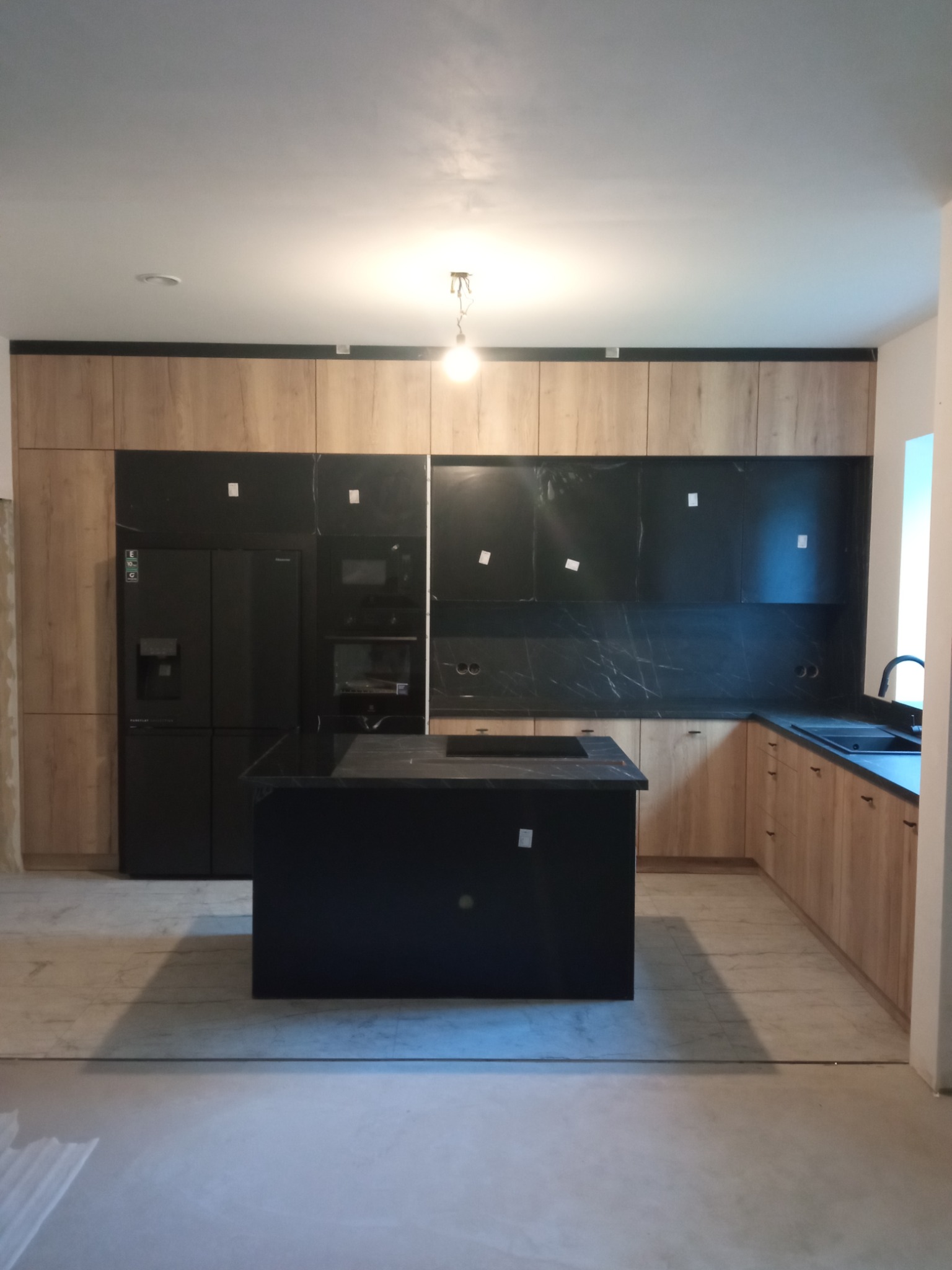 Nowoczesna kuchnia z wyspą: drewniane szafki, czarne blaty i sprzęt AGD. Minimalistyczny design, surowy beton na podłodze, lampa wisząca.